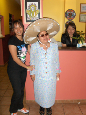 Nanta wears a huge sombrero (hat with a large brim) and stands with Ann in front of the counter with the woman manager smiling.